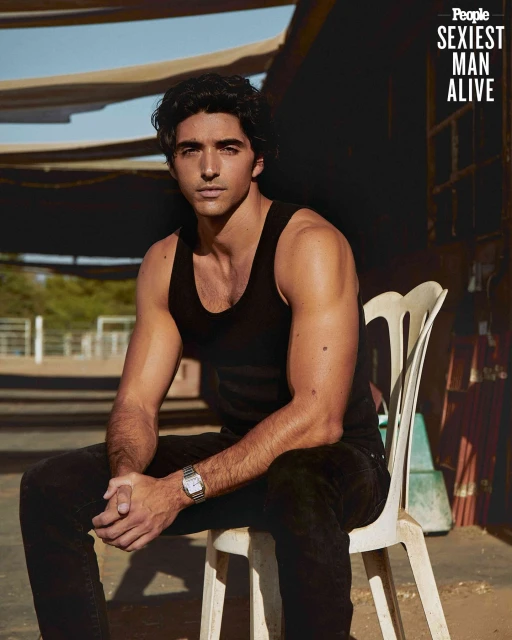 Portrait of a handsome man with dark hair in a black tank top sitting on a white chair outdoors, with a 'Sexiest Man Alive' banner.