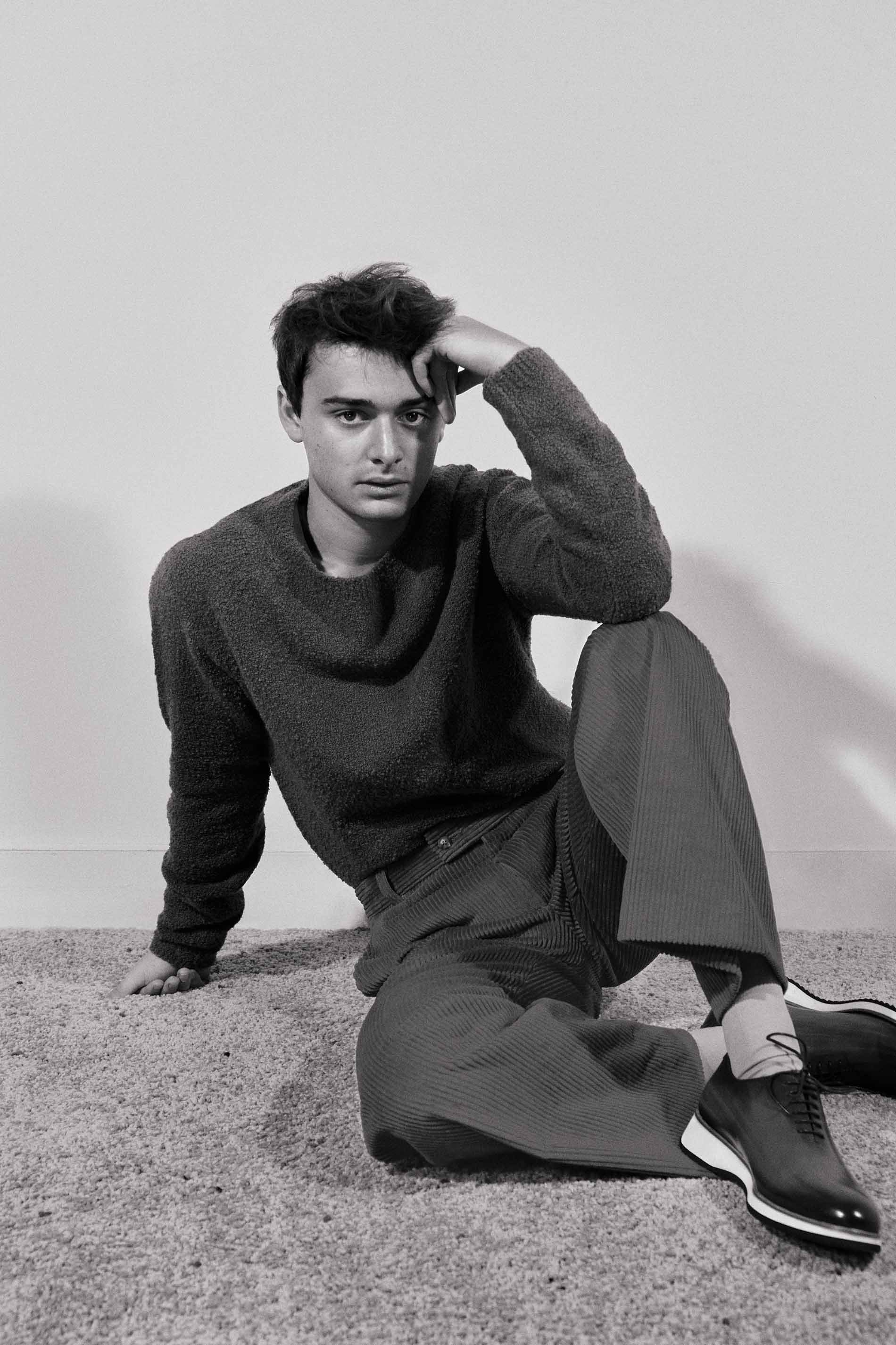 Monochrome portrait of a young man seated on the floor wearing a dark sweater, trousers, and dark lace-up shoes, by Isaac Anthony.