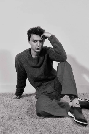 Monochrome portrait of a young man seated on the floor wearing a dark sweater, trousers, and dark lace-up shoes, by Isaac Anthony.