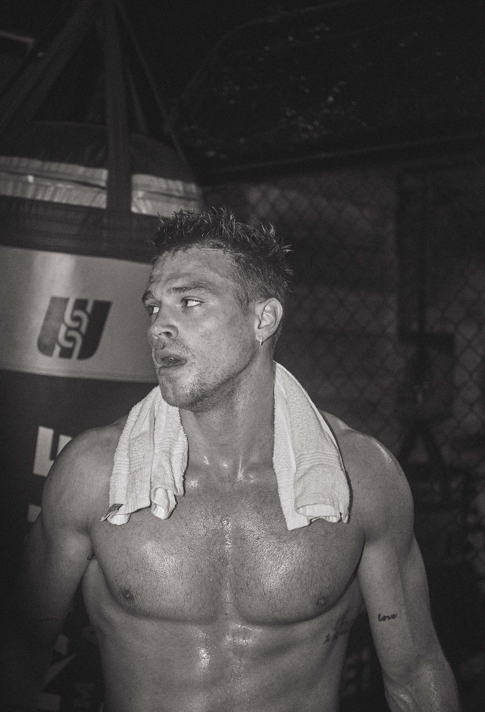 Sweaty shirtless man with a white towel draped over his shoulders in a dark gym with a punching bag visible in the background.