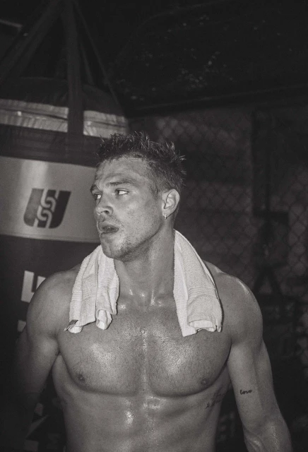 Sweaty shirtless man with a white towel draped over his shoulders in a dark gym with a punching bag visible in the background.