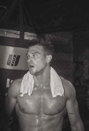 Sweaty shirtless man with a white towel draped over his shoulders in a dark gym with a punching bag visible in the background.