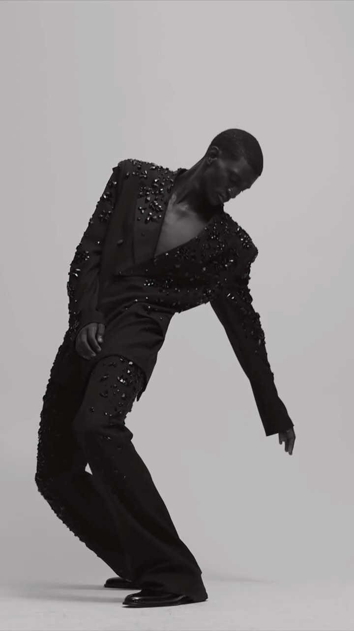 A fashion model in an all-black suit embellished with black stones strikes a dramatic pose in a studio against a neutral background.