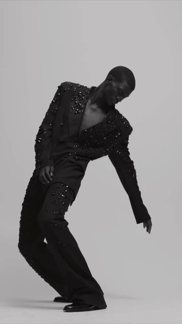 A fashion model in an all-black suit embellished with black stones strikes a dramatic pose in a studio against a neutral background.