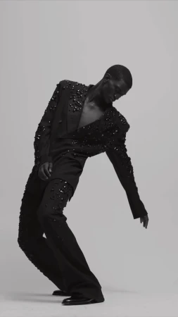 A fashion model in an all-black suit embellished with black stones strikes a dramatic pose in a studio against a neutral background.