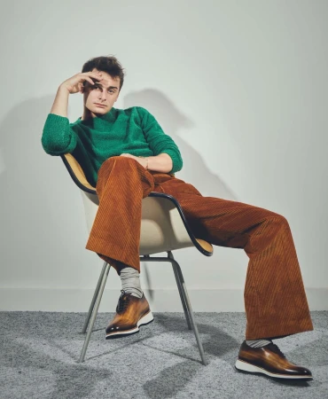 Noah Schnapp wearing a green knit sweater and brown corduroy trousers sitting on a chair against a white wall by Isaac Anthony