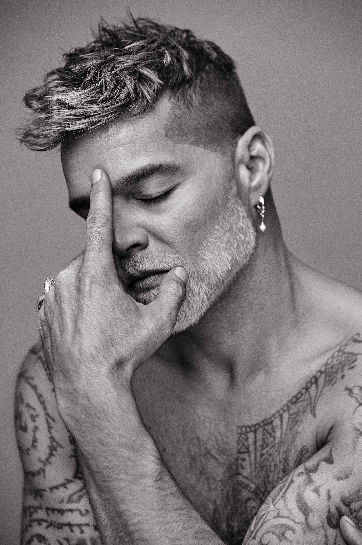 Close-up portrait of a man with salt-and-pepper hair and tattoos, with his hand on his face in a pensive pose against a grey background.