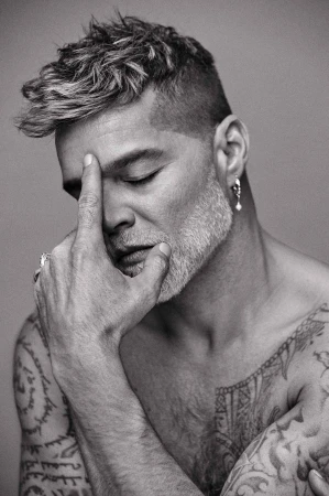 Close-up portrait of a man with salt-and-pepper hair and tattoos, with his hand on his face in a pensive pose against a grey background.