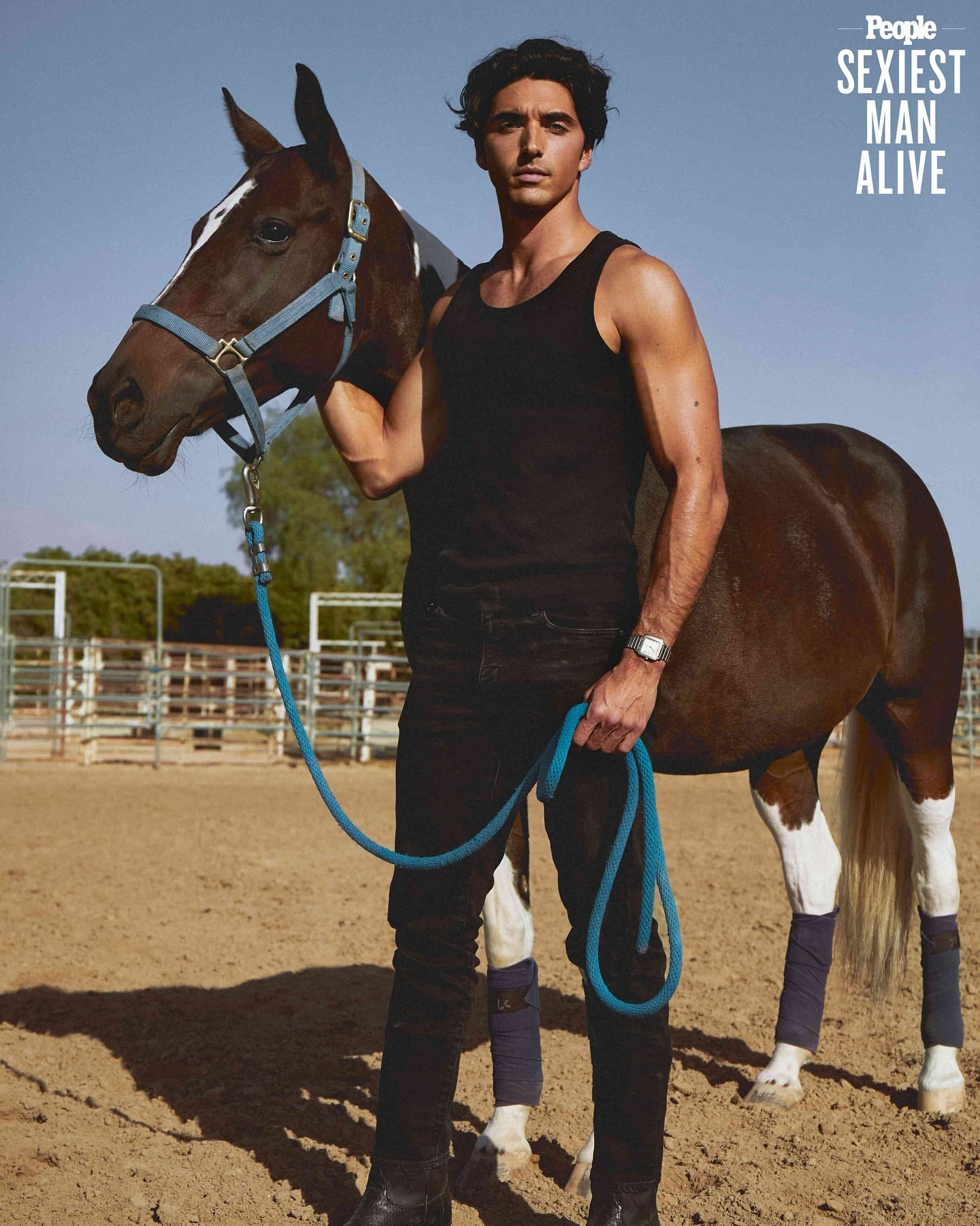Man and horse stand in an outdoor paddock. He wears a tank top and jeans, holding the horse's lead rope. Bright daylight.
