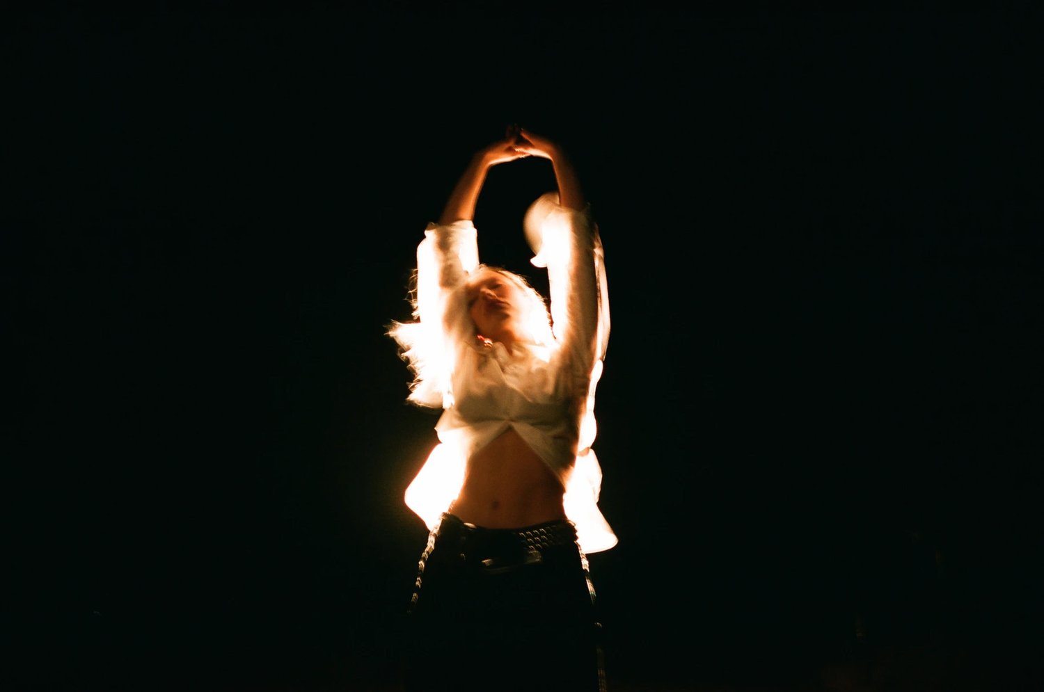 A person in a white buttoned shirt with arms raised high against a dark background, lit by strong backlighting.
