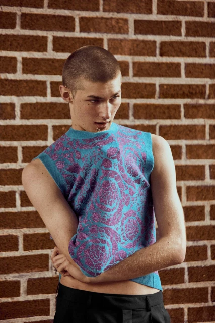 A young man poses against a brick wall, wearing a stylish turquoise and purple patterned tank top and black dress pants.