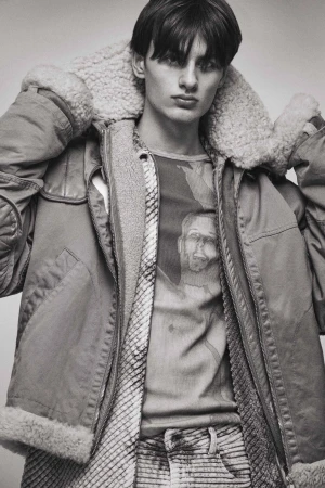 Monochrome portrait of a young male model wearing a heavy shearling-lined jacket and patterned t-shirt by Isaac Anthony.