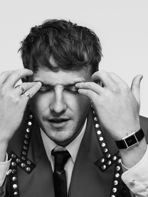 Black and white portrait of a man with hands on his face, wearing a suit jacket with metal studs, against a plain white backdrop.