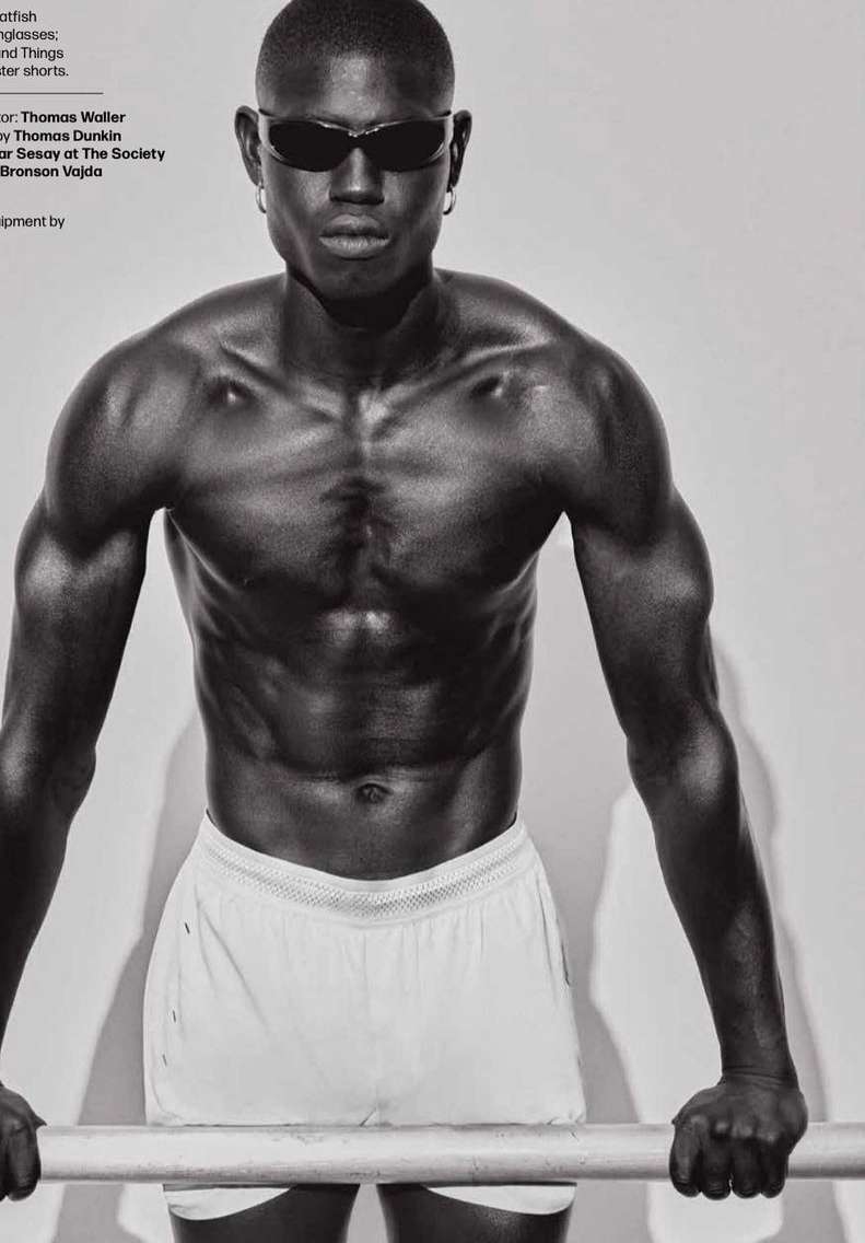 Athletic African American man with sunglasses and white shorts poses with a metal bar in a black and white fashion shot.