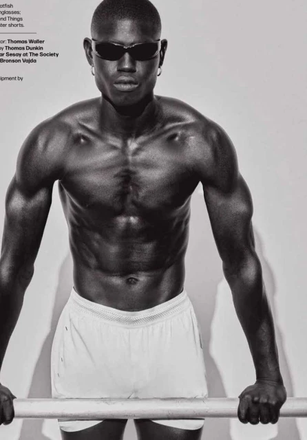 Athletic African American man with sunglasses and white shorts poses with a metal bar in a black and white fashion shot.