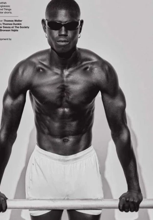 Athletic African American man with sunglasses and white shorts poses with a metal bar in a black and white fashion shot.