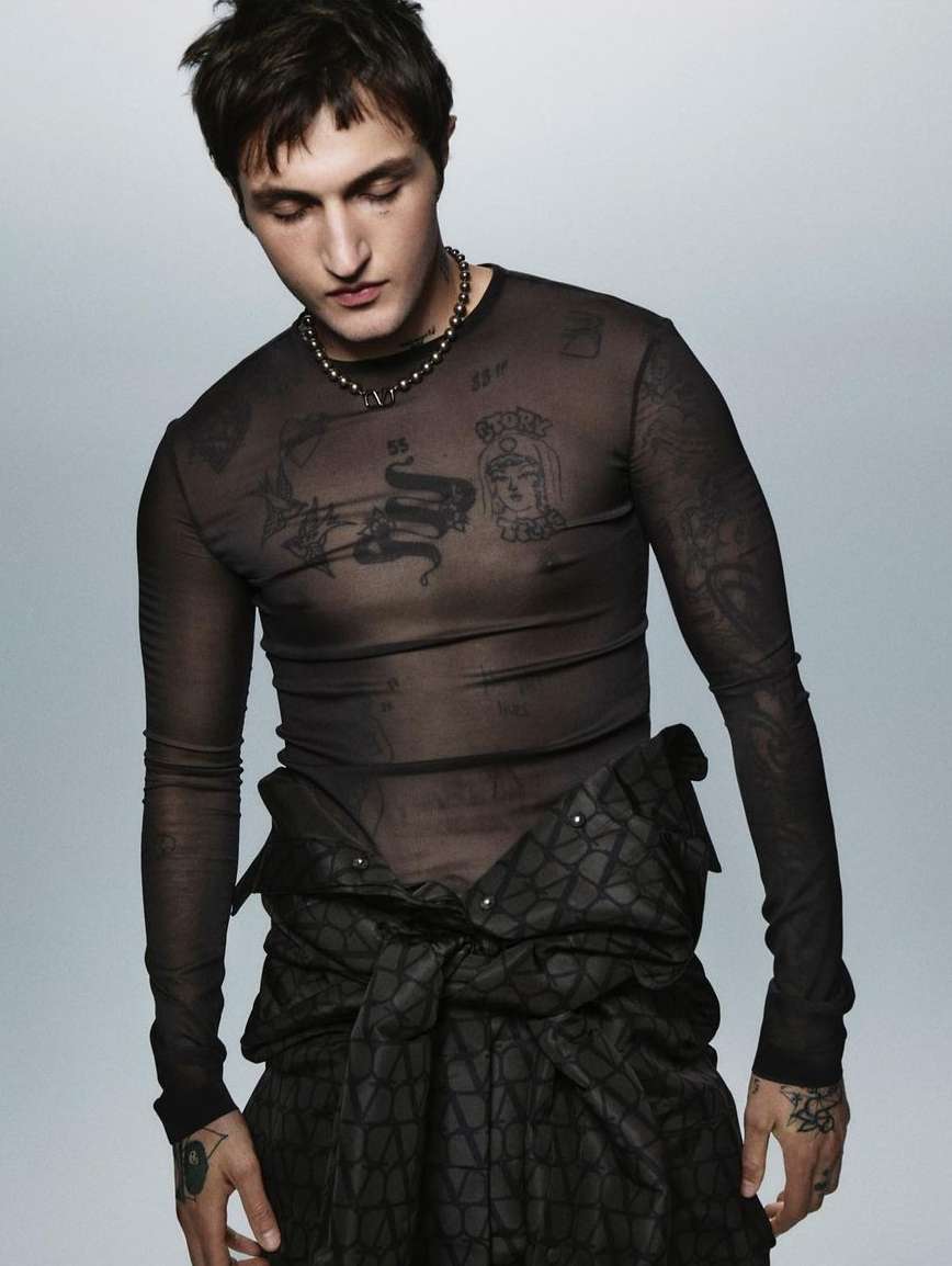 An adult male poses in a dark mesh shirt with tattoos visible on his arms and chest, and a black patterned garment around his waist.