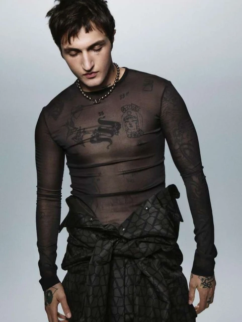 An adult male poses in a dark mesh shirt with tattoos visible on his arms and chest, and a black patterned garment around his waist.