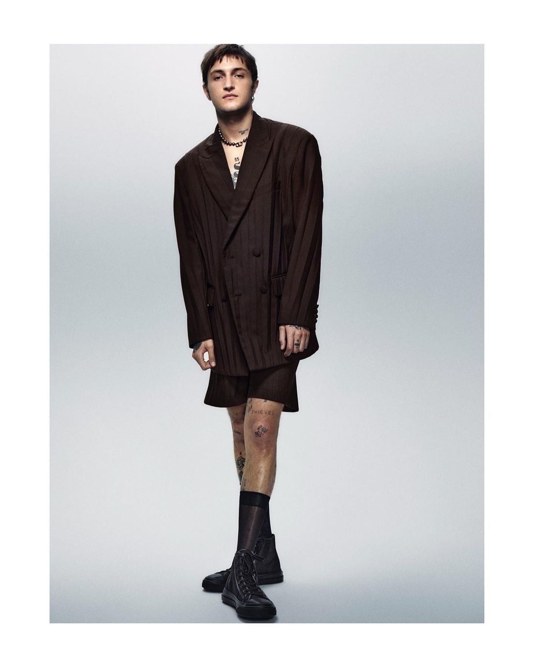 Young adult man in oversized brown suit and black boots standing in a photo studio, tattoos visible on arms and legs, fashion portrait.