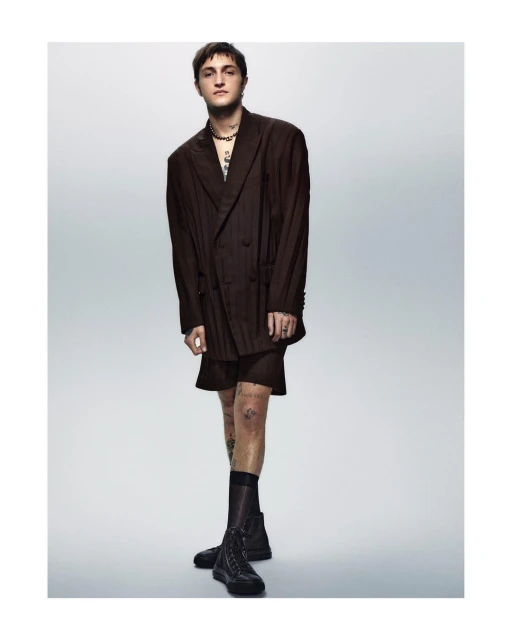 Young adult man in oversized brown suit and black boots standing in a photo studio, tattoos visible on arms and legs, fashion portrait.