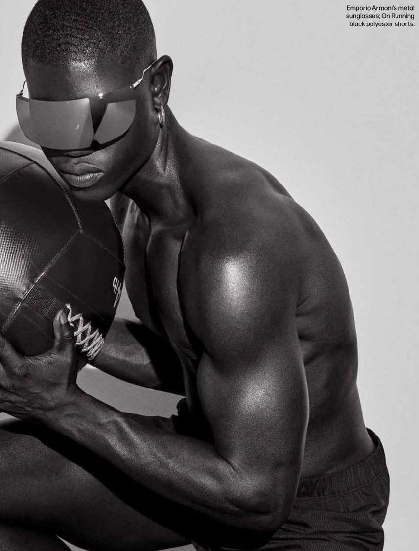 Athletic dark-skinned male model poses shirtless in black shorts and sunglasses, holding a black medicine ball against a grey background.