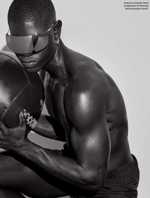 Athletic dark-skinned male model poses shirtless in black shorts and sunglasses, holding a black medicine ball against a grey background.