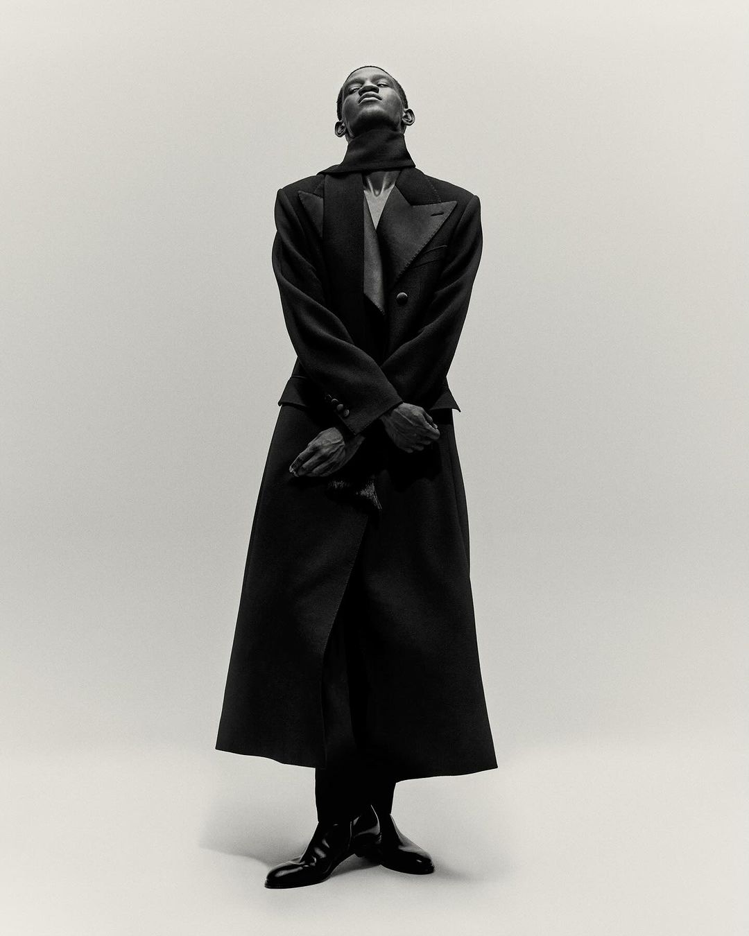 An elegant Black male fashion model poses in a tailored black coat and suit, against a clean background in a portrait fashion shot.
