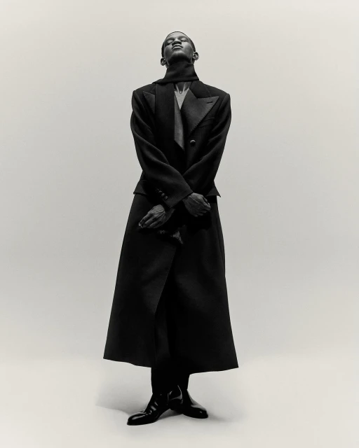 An elegant Black male fashion model poses in a tailored black coat and suit, against a clean background in a portrait fashion shot.