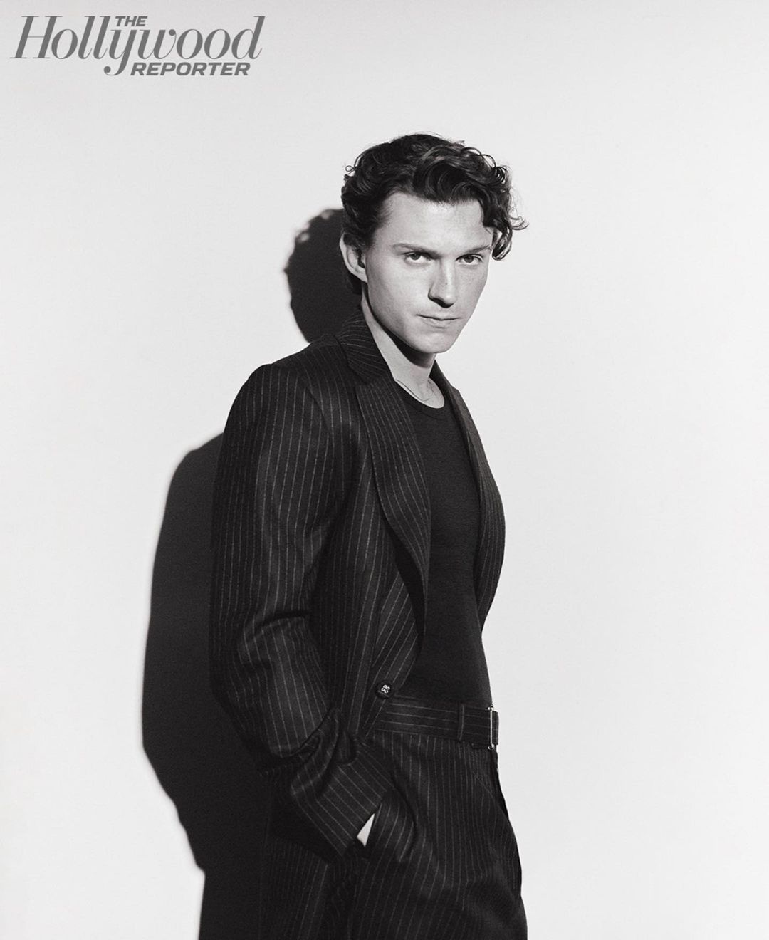 Black and white portrait of a young adult male in a pinstripe suit jacket and dark shirt, posing against a white backdrop in a photo studio.