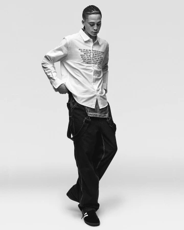 Young man wearing a white button-down shirt with printed text and dark overalls against a plain white background.