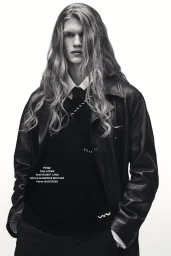 Black and white portrait of a male model with long wavy hair wearing a leather coat, vest, and tie against a plain white background.