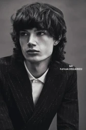 Close-up portrait of a young man with dark shaggy hair wearing a pinstripe suit jacket and white collared shirt against grey.
