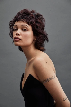 Portrait of a woman with dark curly hair and a black strapless top against a grey seamless studio background.