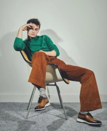Noah Schnapp wearing a green knit sweater and brown corduroy trousers sitting on a chair against a white wall by Isaac Anthony