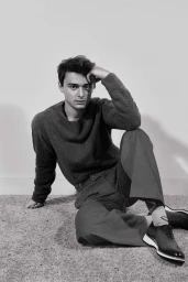 Monochrome portrait of a young man seated on the floor wearing a dark sweater, trousers, and dark lace-up shoes, by Isaac Anthony.