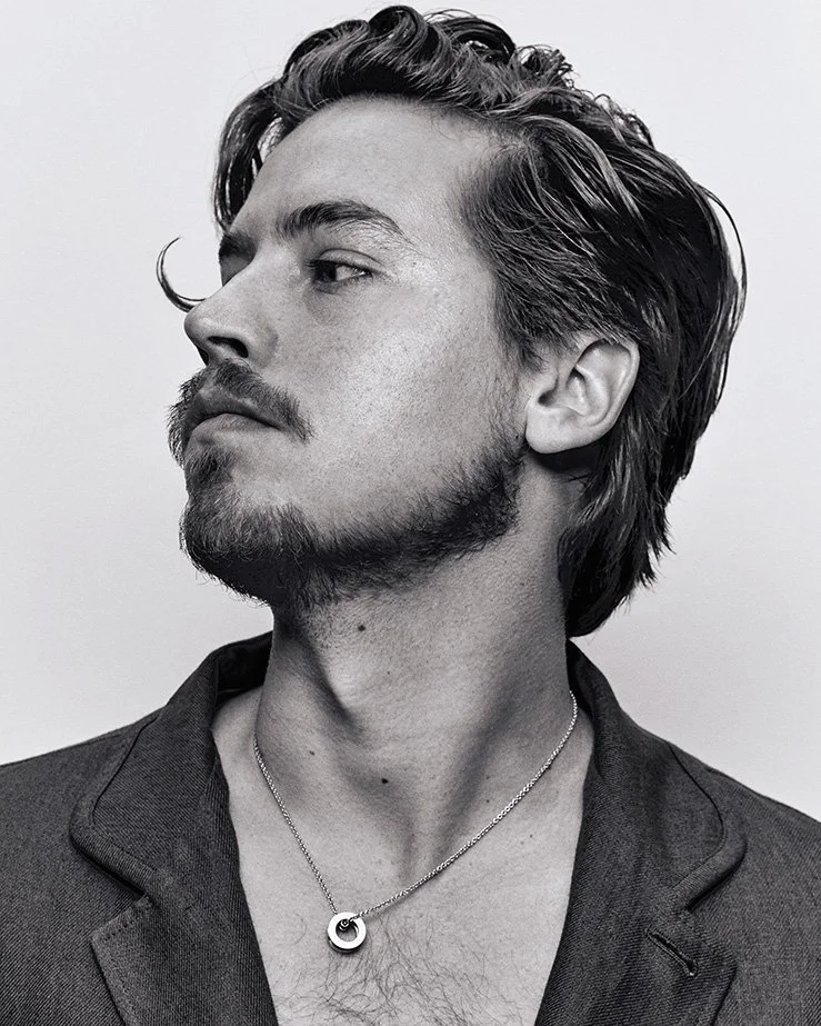 Black and white portrait of a man with wavy hair and facial hair wearing a necklace against a light background