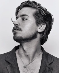 Black and white portrait of a man with wavy hair and facial hair wearing a necklace against a light background