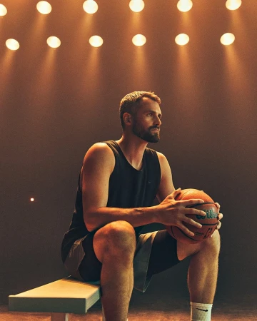 Athlete sitting on a bench holding a basketball under warm overhead lights by Isaac Anthony