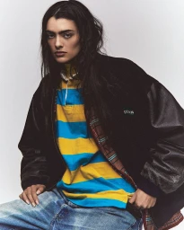 Model with long dark hair wearing a striped yellow and blue polo shirt under a black leather jacket and denim jeans.