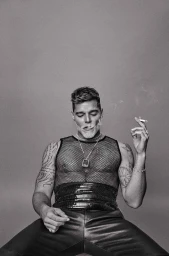 Black and white portrait of a man wearing a mesh tank top and holding a cigarette against a plain background