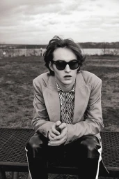 Young man in sunglasses and blazer seated outdoors on a bench, photographed by Isaac Anthony in monochrome.