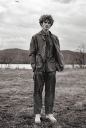 Monochrome portrait of a young man with curly hair wearing a jacket and trousers standing outdoors near a lake.