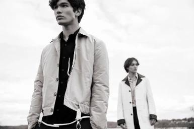 Two young male models in jackets outdoors under an overcast sky, photographed by Isaac Anthony in black and white.