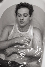 Monochrome portrait of a young man wearing a wet white tank top while lying inside a bathtub.