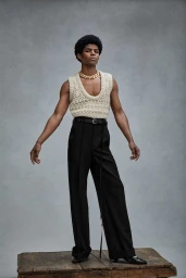 Black man wearing a cream knit vest, black trousers, and a chunky gold necklace standing on a wooden box
