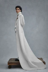 Model wearing a long white coat with a train standing on a weathered wooden platform against a grey textured studio backdrop.