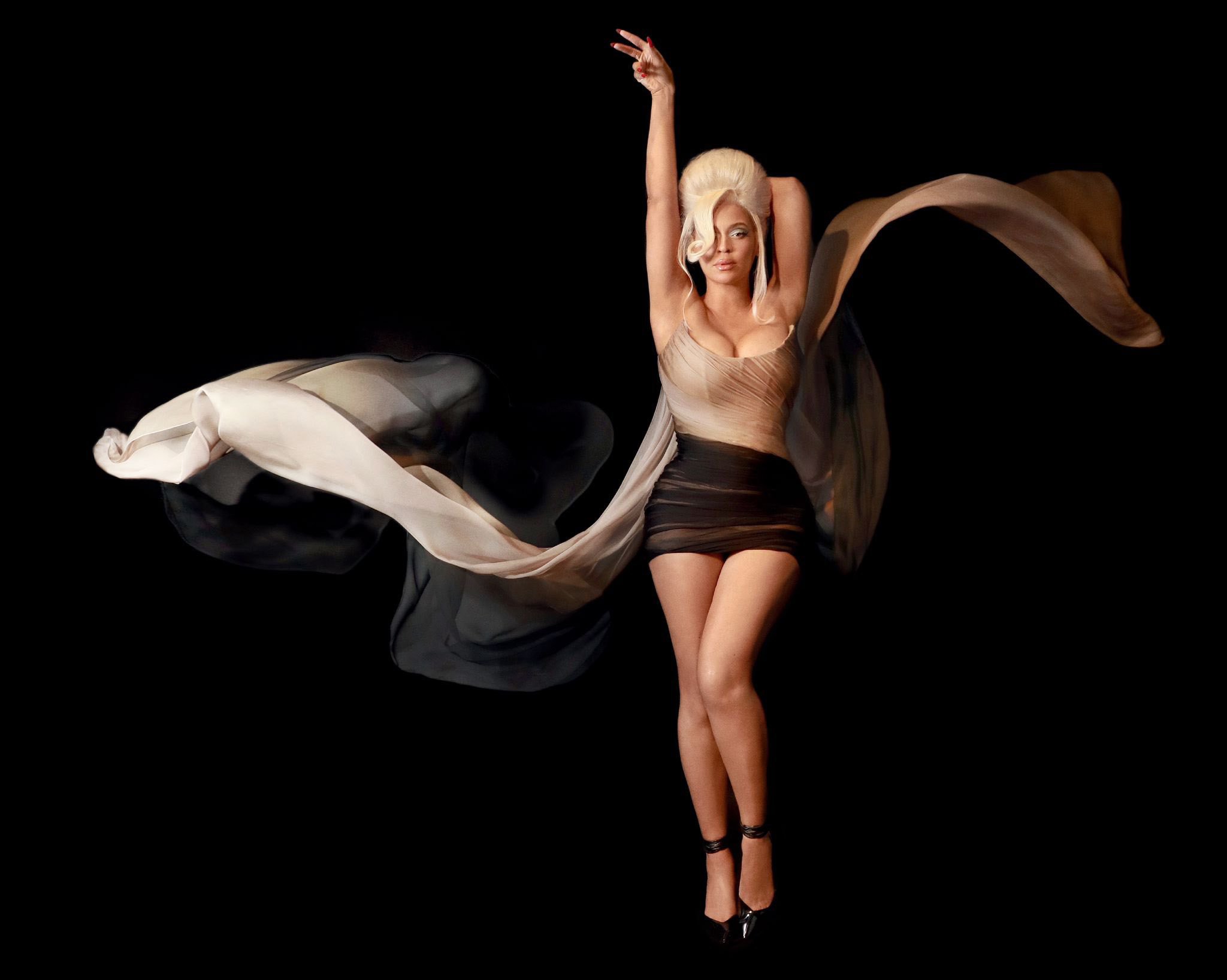 Woman in flowing beige dress poses in a black studio, one arm raised, sheer fabric flowing dramatically around her against the dark background.