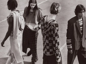 Four male models in casual knitwear and outerwear walking on an outdoor asphalt surface in a black and white editorial photograph.