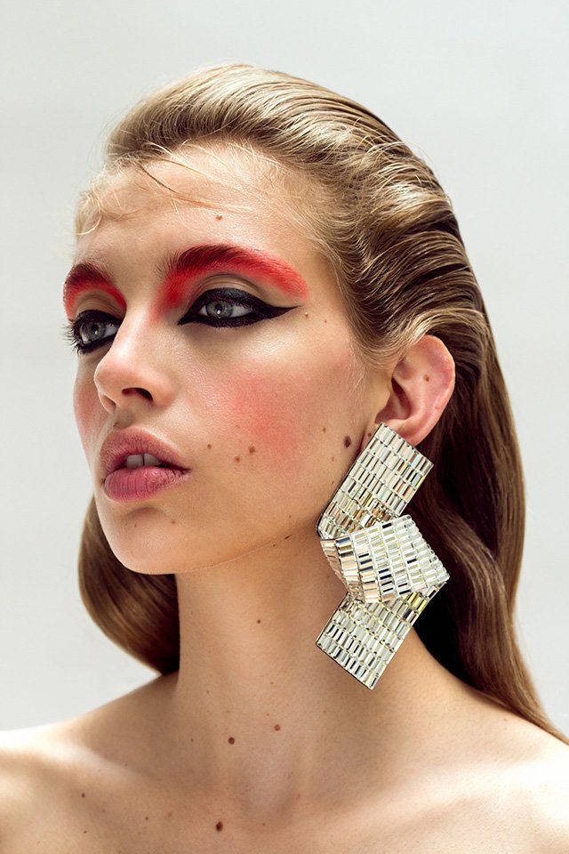 Headshot of a fair-skinned woman with wet-look blonde hair, dramatic red and black eye makeup, and large geometric silver earrings.