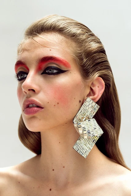 Headshot of a fair-skinned woman with wet-look blonde hair, dramatic red and black eye makeup, and large geometric silver earrings.